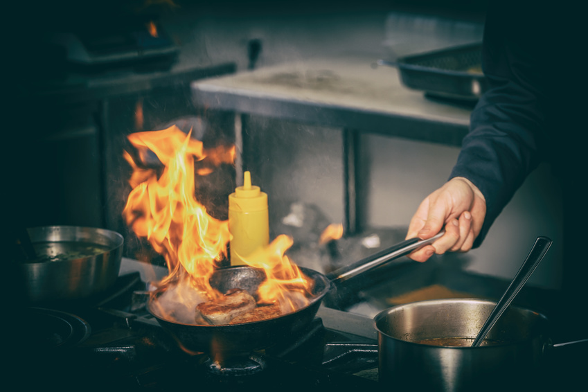 A person is cooking a meat with a fire blazing on the pan that may cause a burn injury.