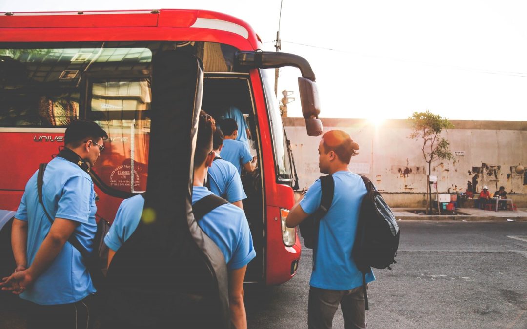 Group of men wearing blue shirt with bags and are lining up and getting inside of a red bus to avoid bus accidents involving best serious injury attorney with sunrise as a background.